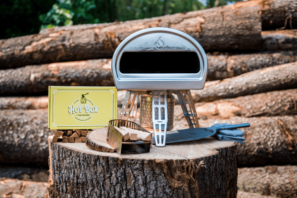 A box of Hot Box Cooking wood sits on top of a log and next to a portable pizza oven