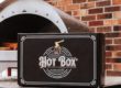 A Cru pizza oven next to a box of Hot Box Cooking Wood