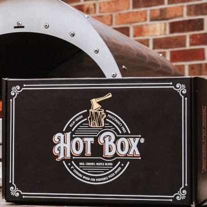 A Cru pizza oven next to a box of Hot Box Cooking Wood